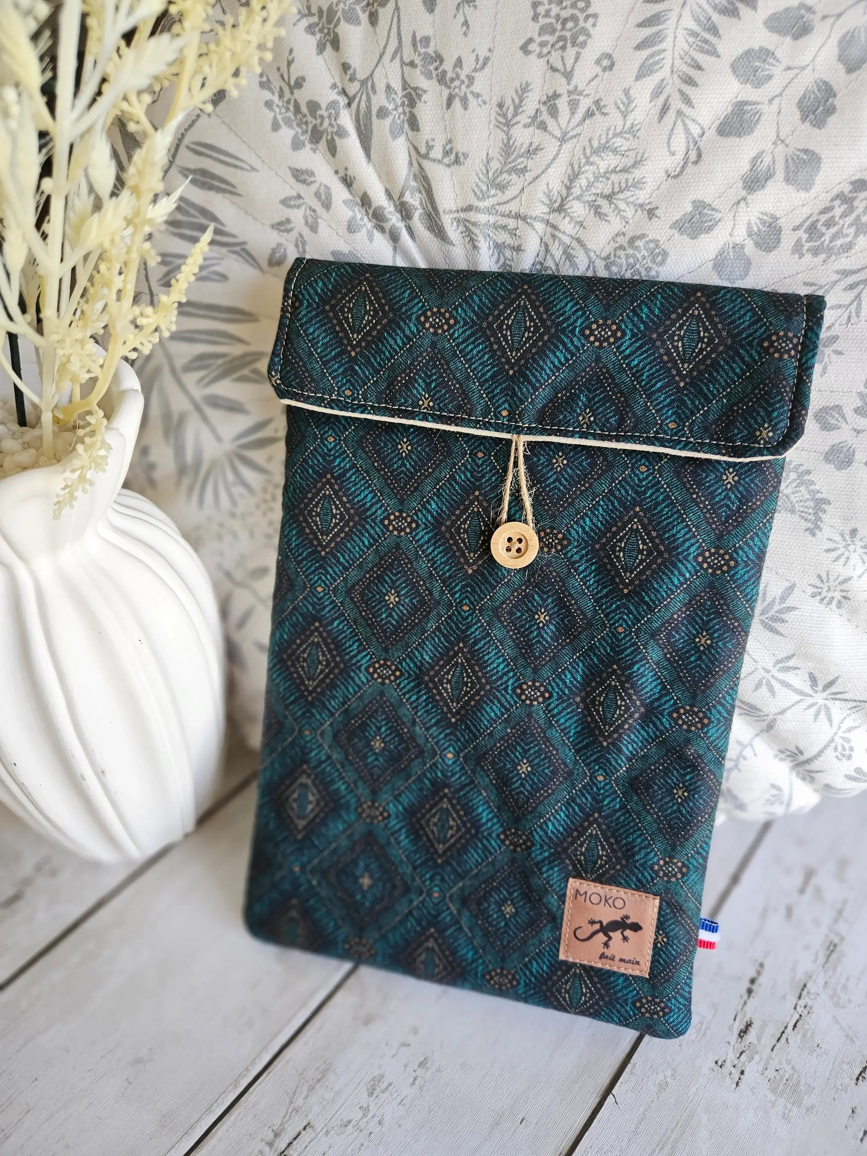 Pochette tissu bleu motif géométrique, bouton bois, sur fond végétal décoratif.