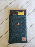 Pochette textile bleue à motif géométrique, bouton bois, contenant un livre, sur fond bois.
