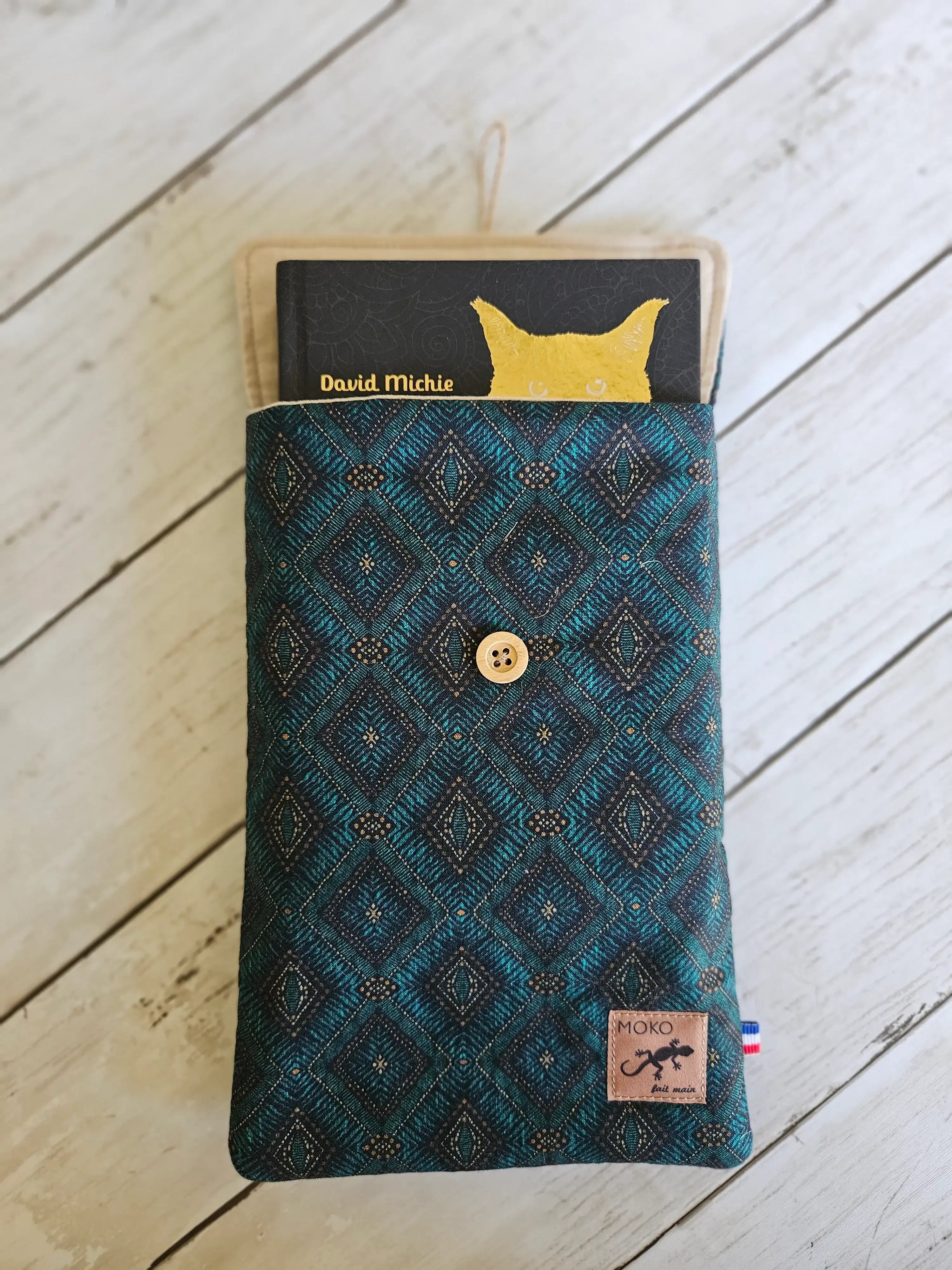 Pochette textile bleue à motif géométrique, bouton bois, contenant un livre, sur fond bois.