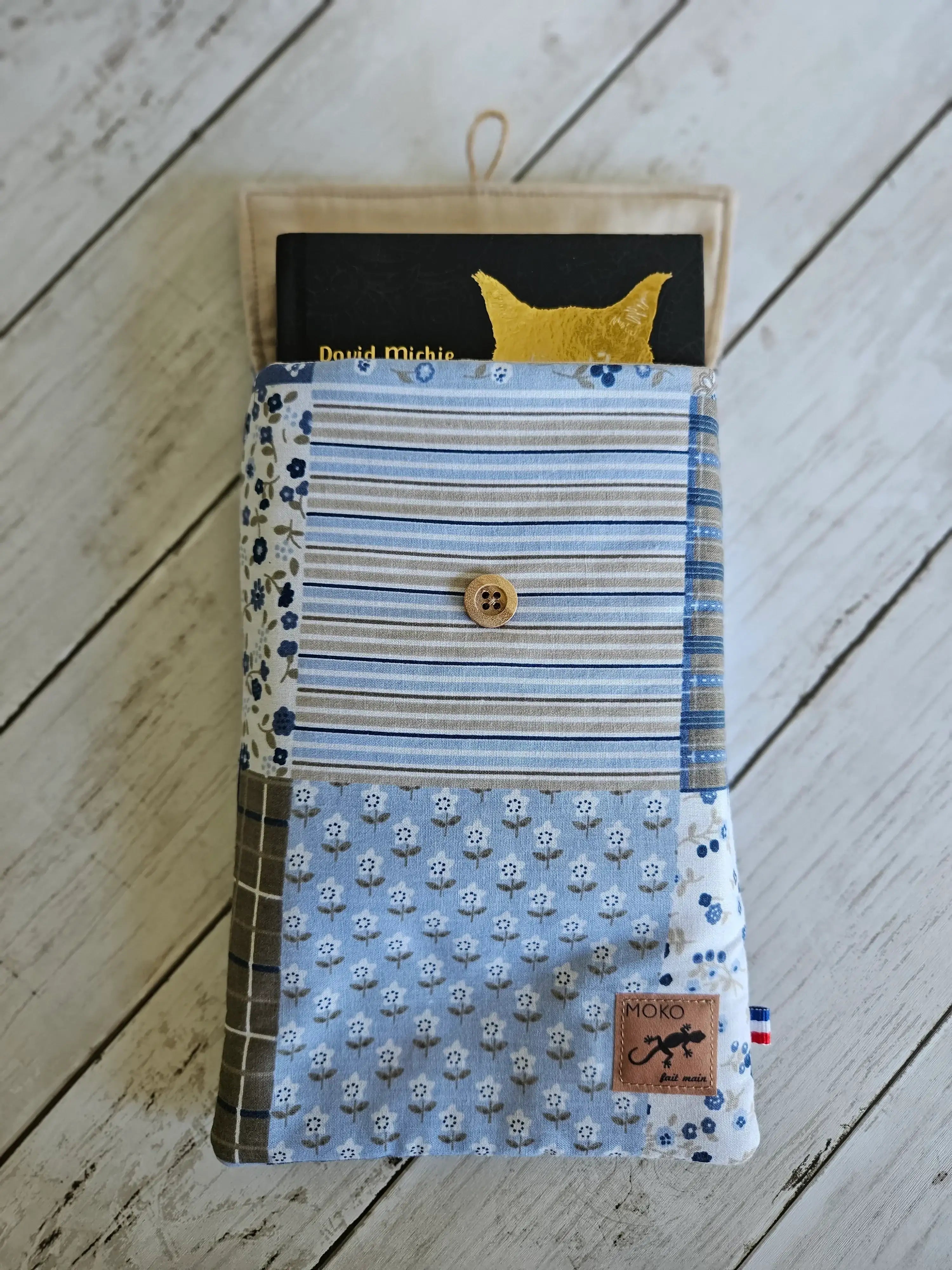 Pochette livre patchwork bleu à motifs floraux et rayures, bouton bois, sur fond bois clair.