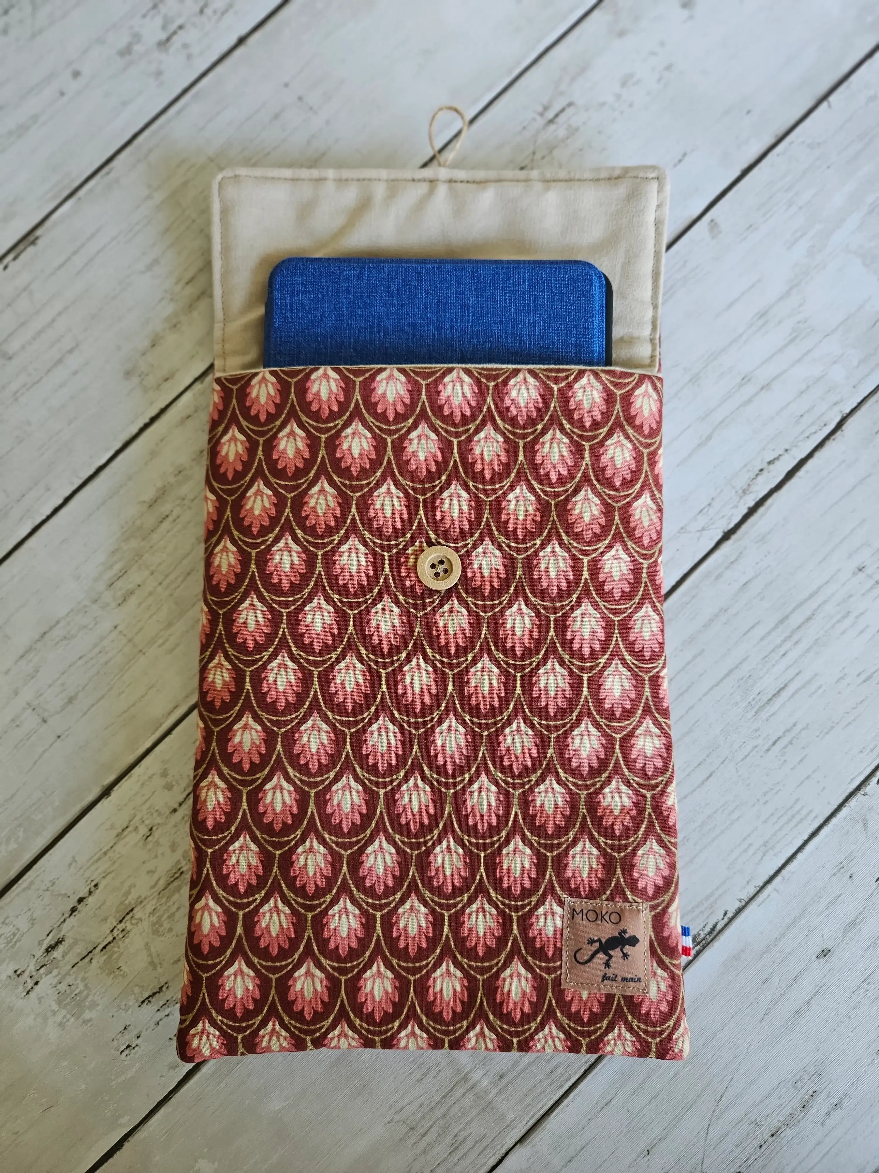 Pochette tissu motif rose avec bouton en bois, contenant une tablette bleue, sur fond bois.