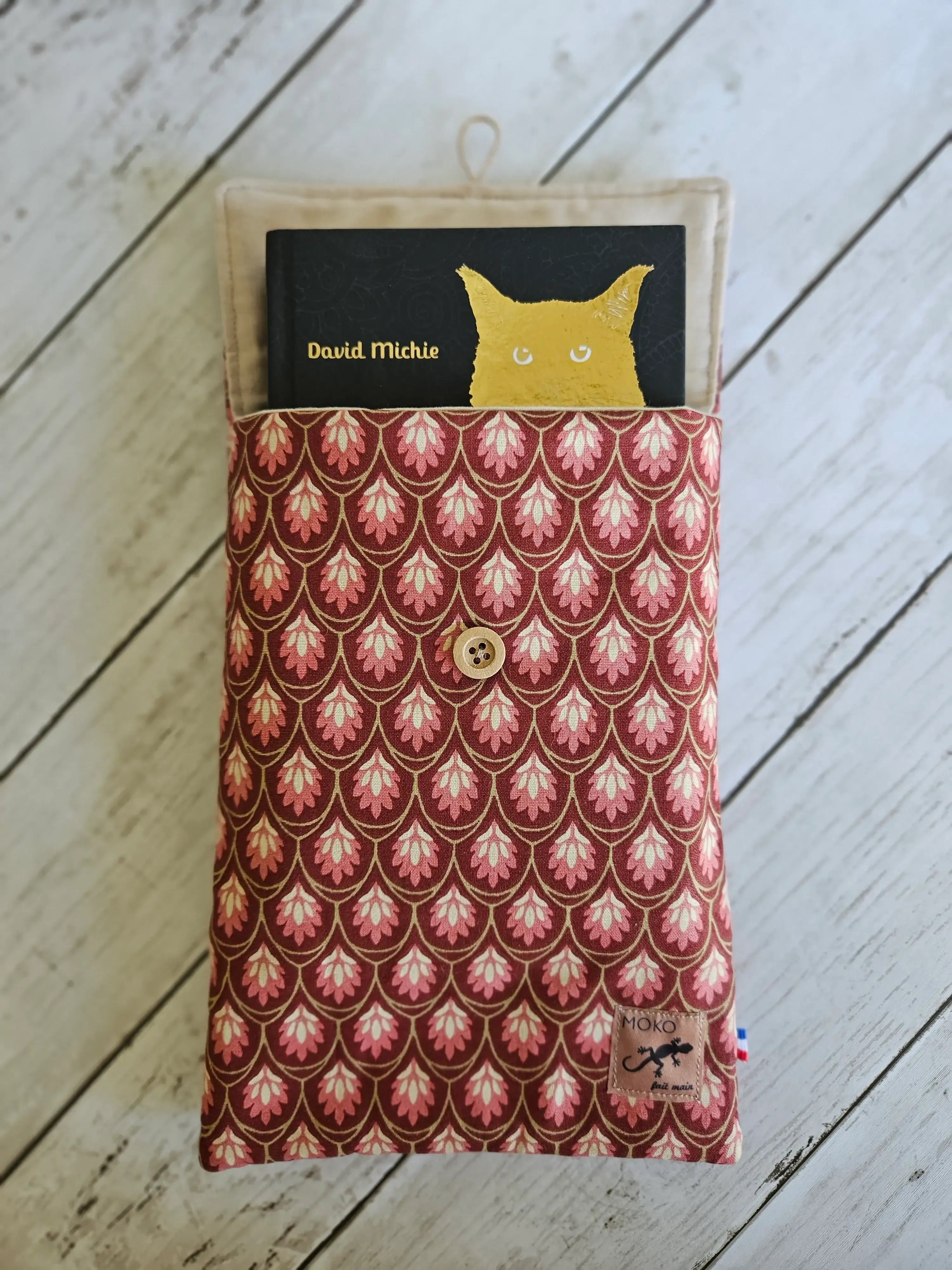 Pochette en tissu imprimé rouge rose avec bouton, contenant un livre, sur fond bois blanc.