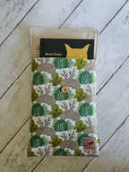 Pochette livre tissu motif forêt vert beige avec bouton bois sur fond en bois blanc, accessoire mode