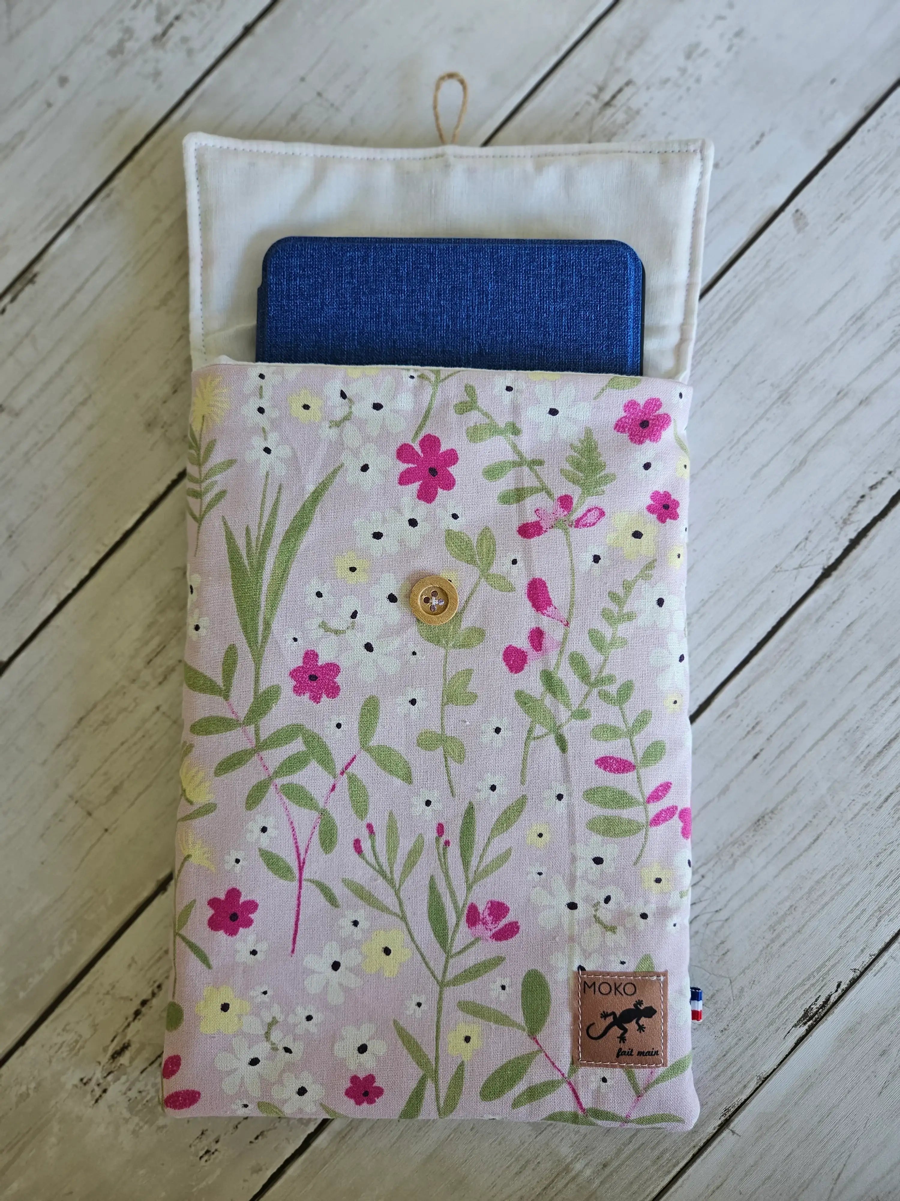 Housse de livre liseuse motif fleuri pastel, bouton bois, sur fond en bois blanc.