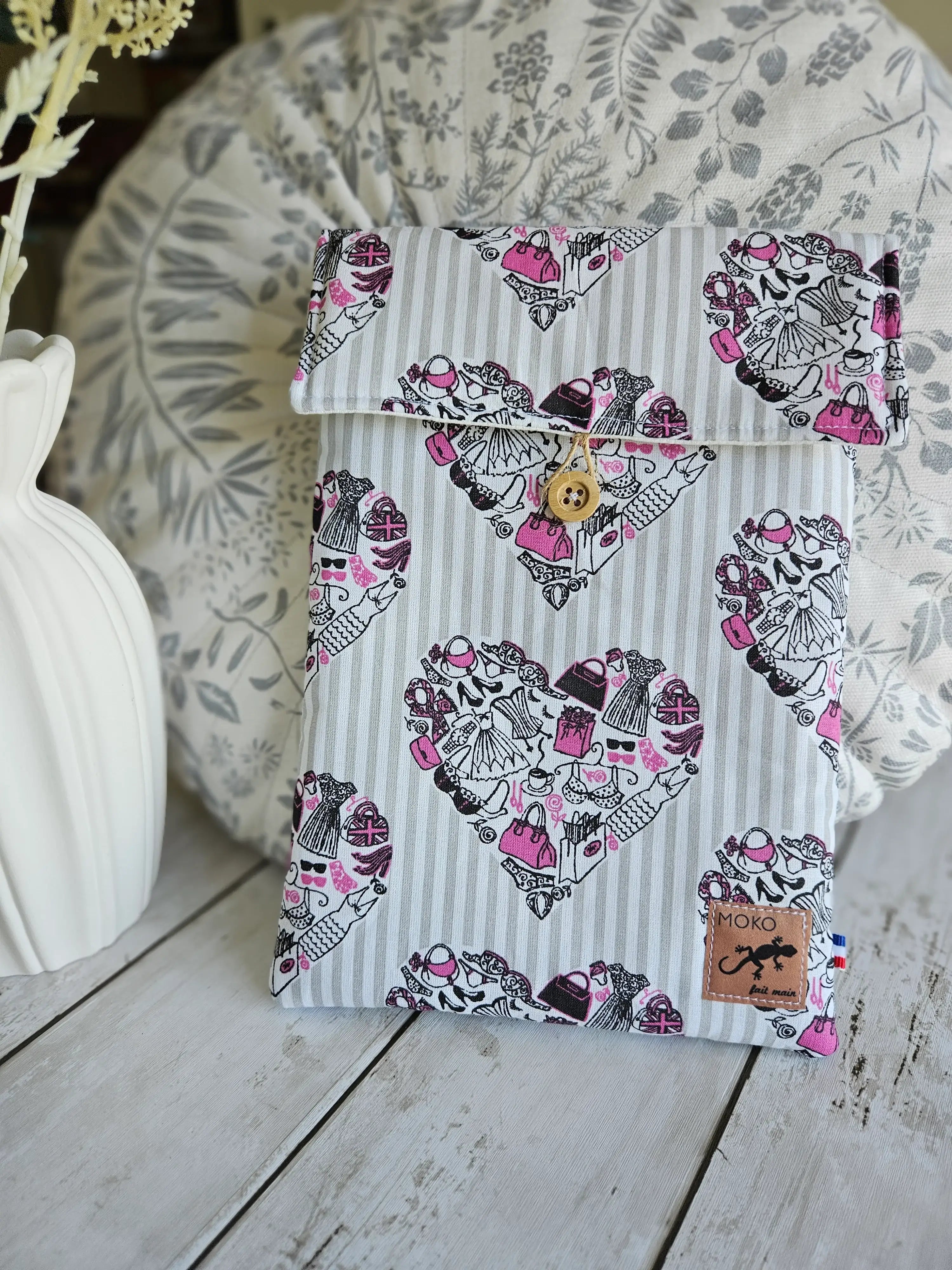 Housse à livre liseuse en tissu à motifs cœurs, fermée par un bouton, sur fond décoratif.
