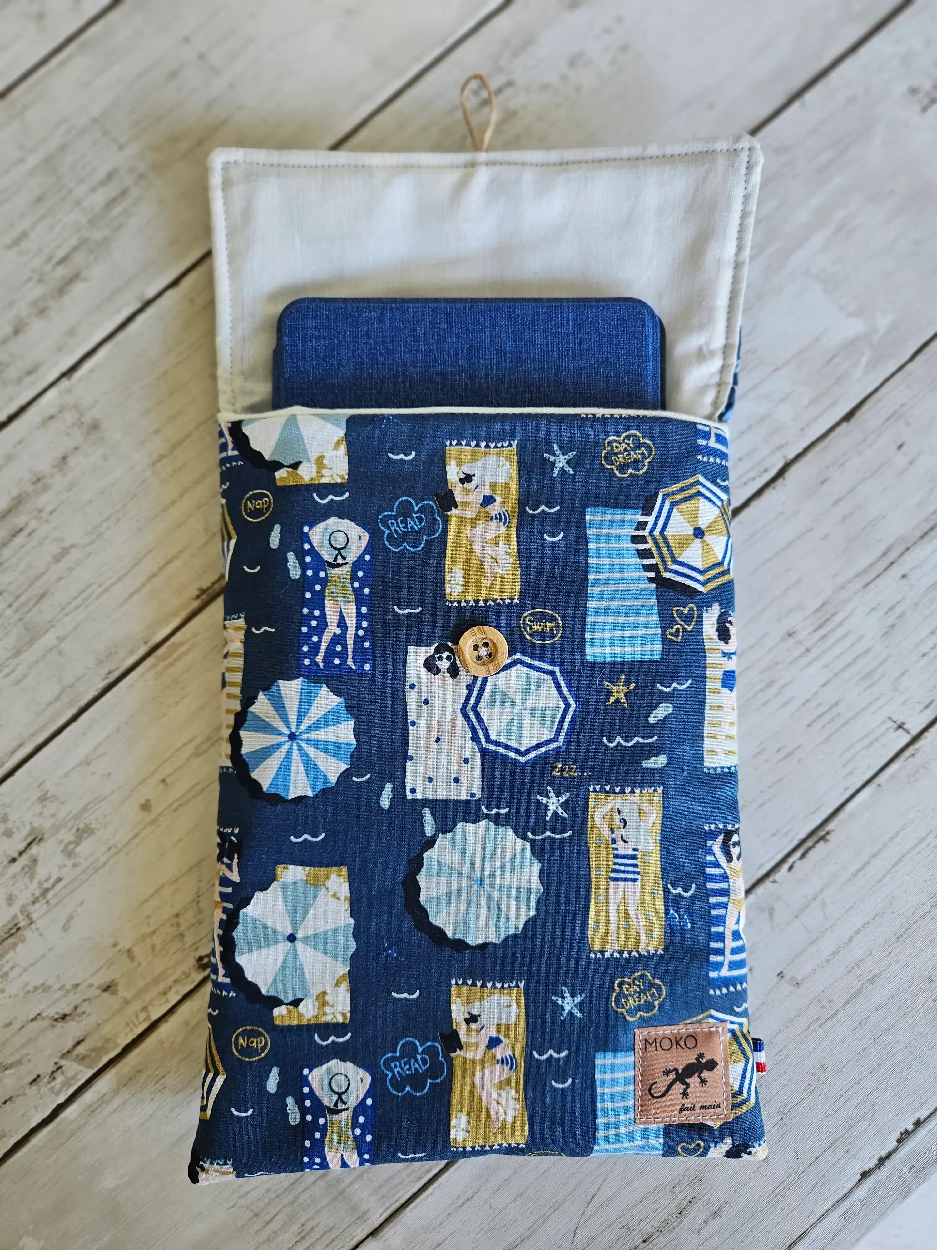 Étui tissu bleu à motifs plage, pour tablette, sur fond bois blanc.
