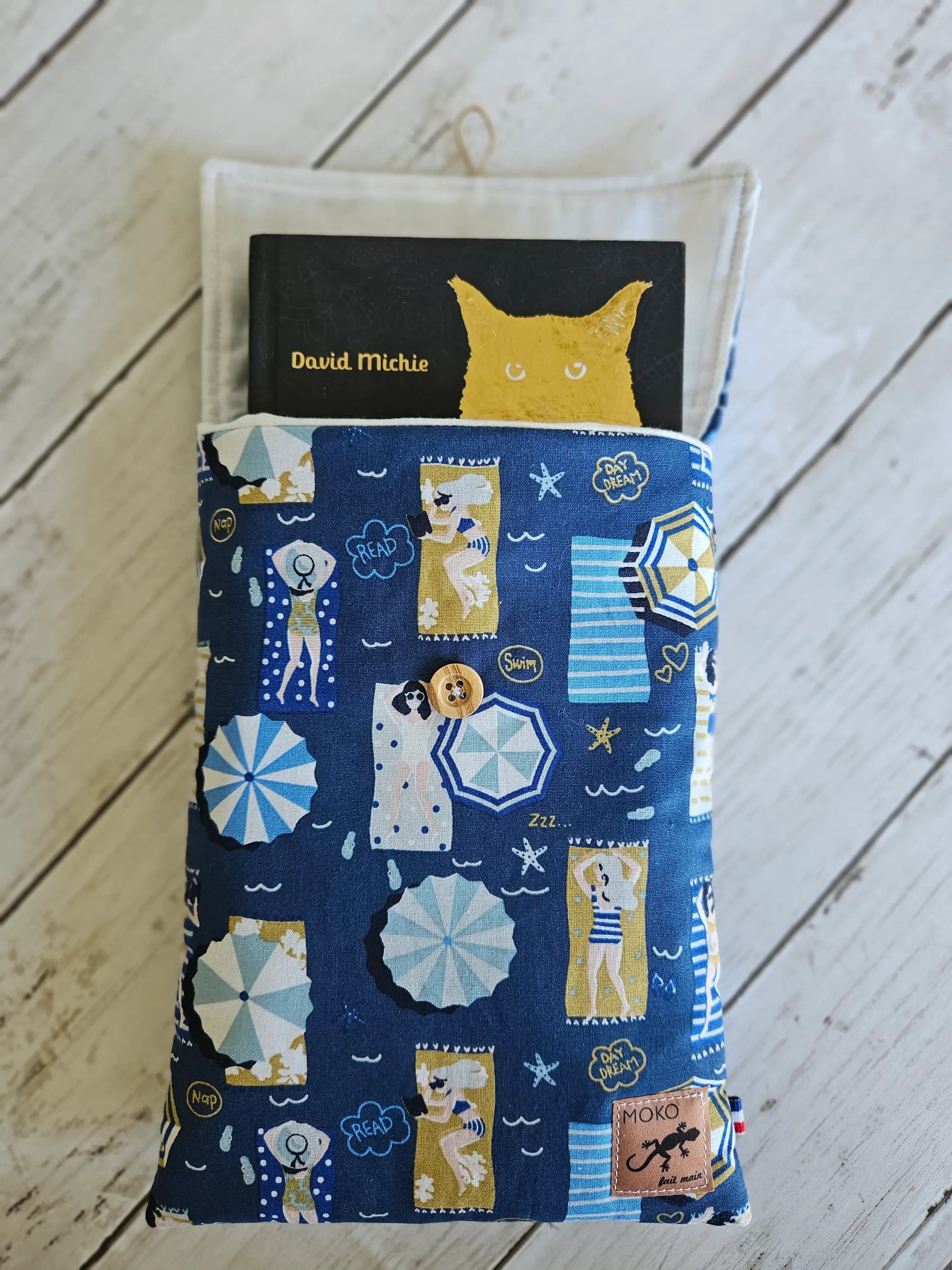 Housse à livre tissu bleu motifs plage, livre inséré, fond bois clair, accessoire lecture.