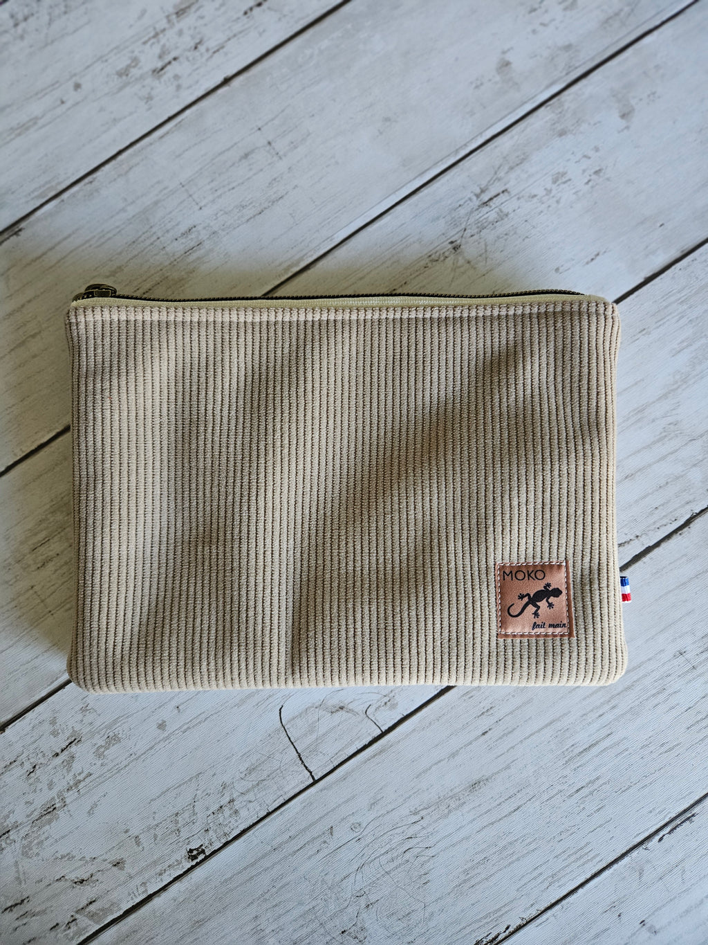 Trousse plate en velours beige côtelé avec zip, étiquette cuir, posée sur bois clair