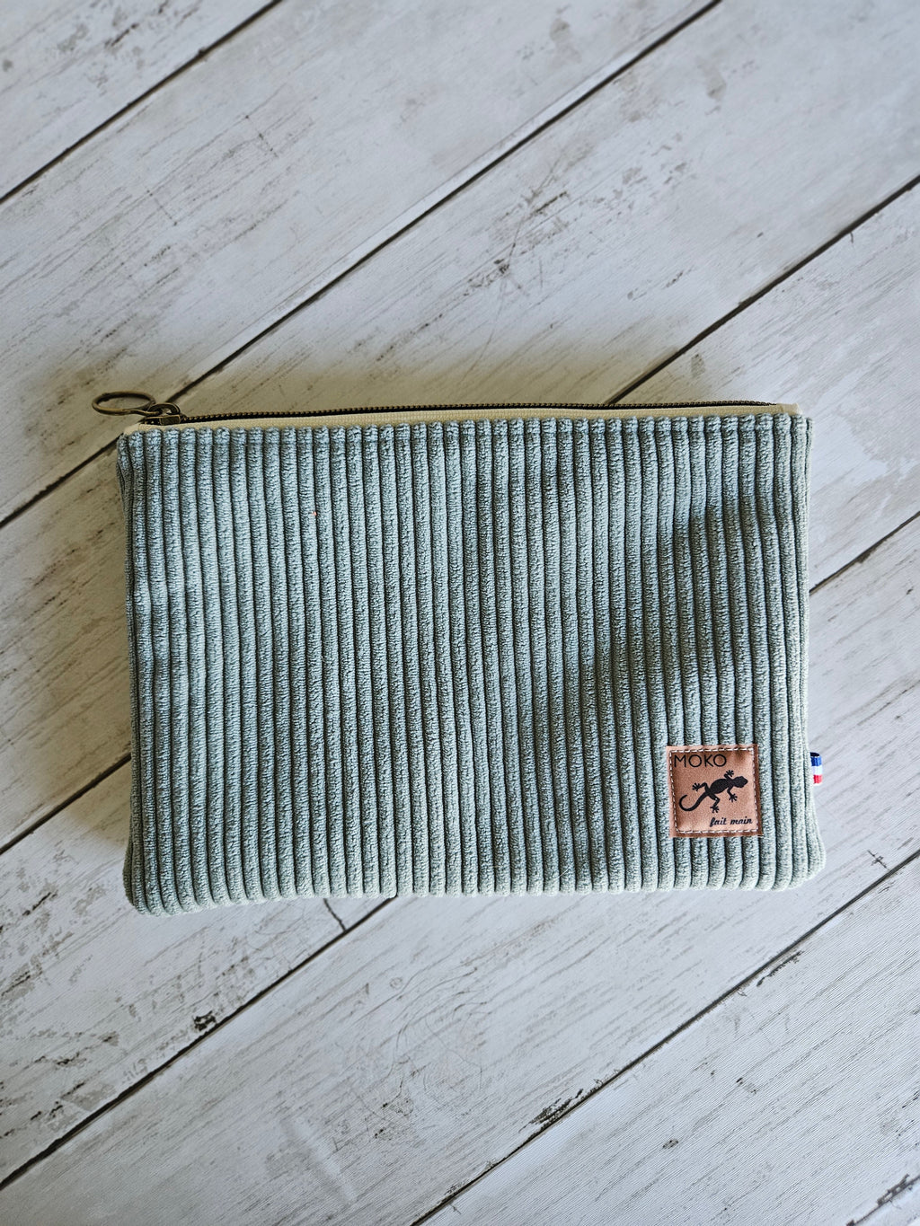 Trousse plate en velours vert d’eau avec fermeture éclair et étiquette cousue sur fond bois.