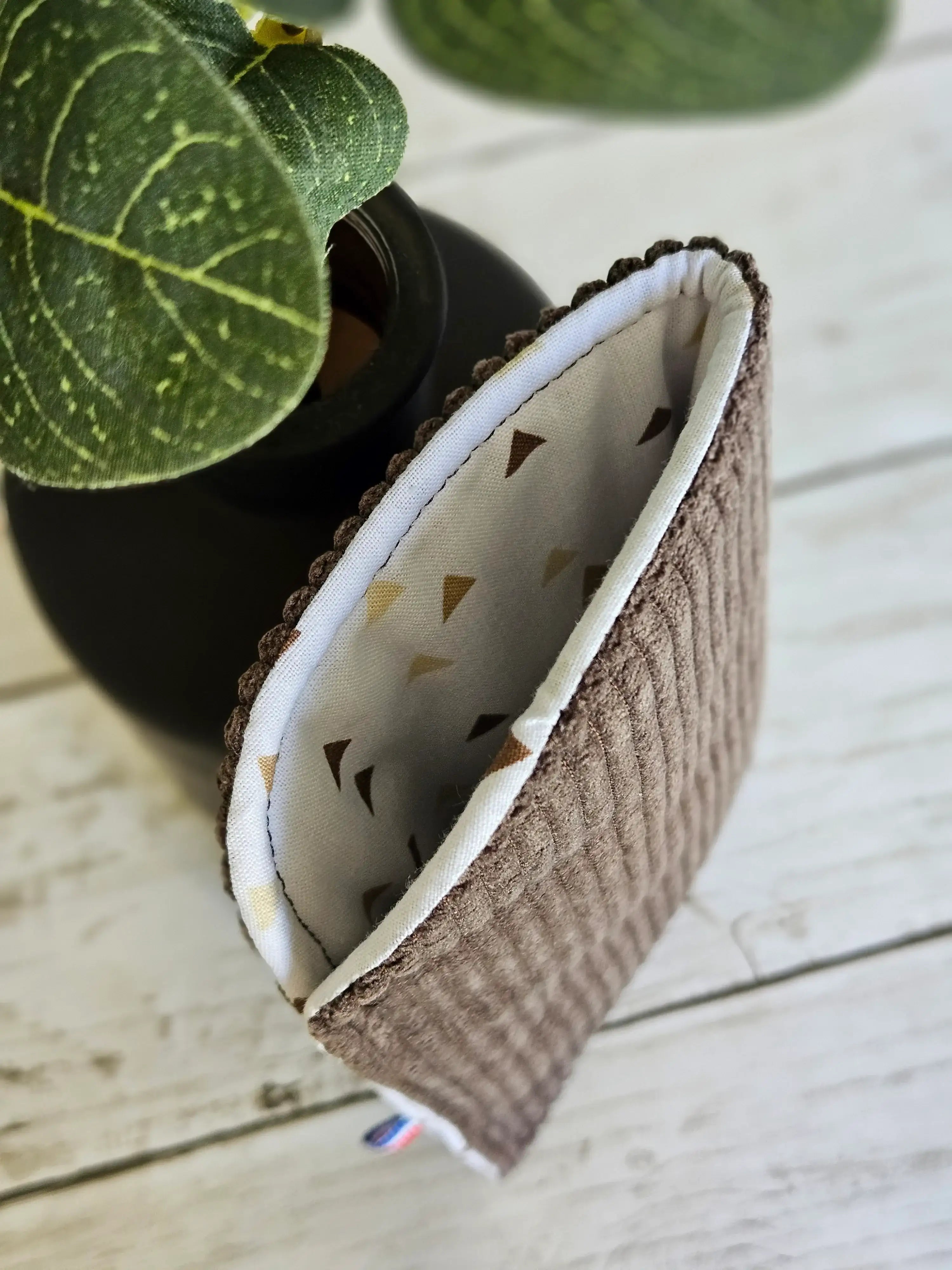 Pochette en tissu côtelé marron, doublure à motifs géométriques, posée près d'une plante.
