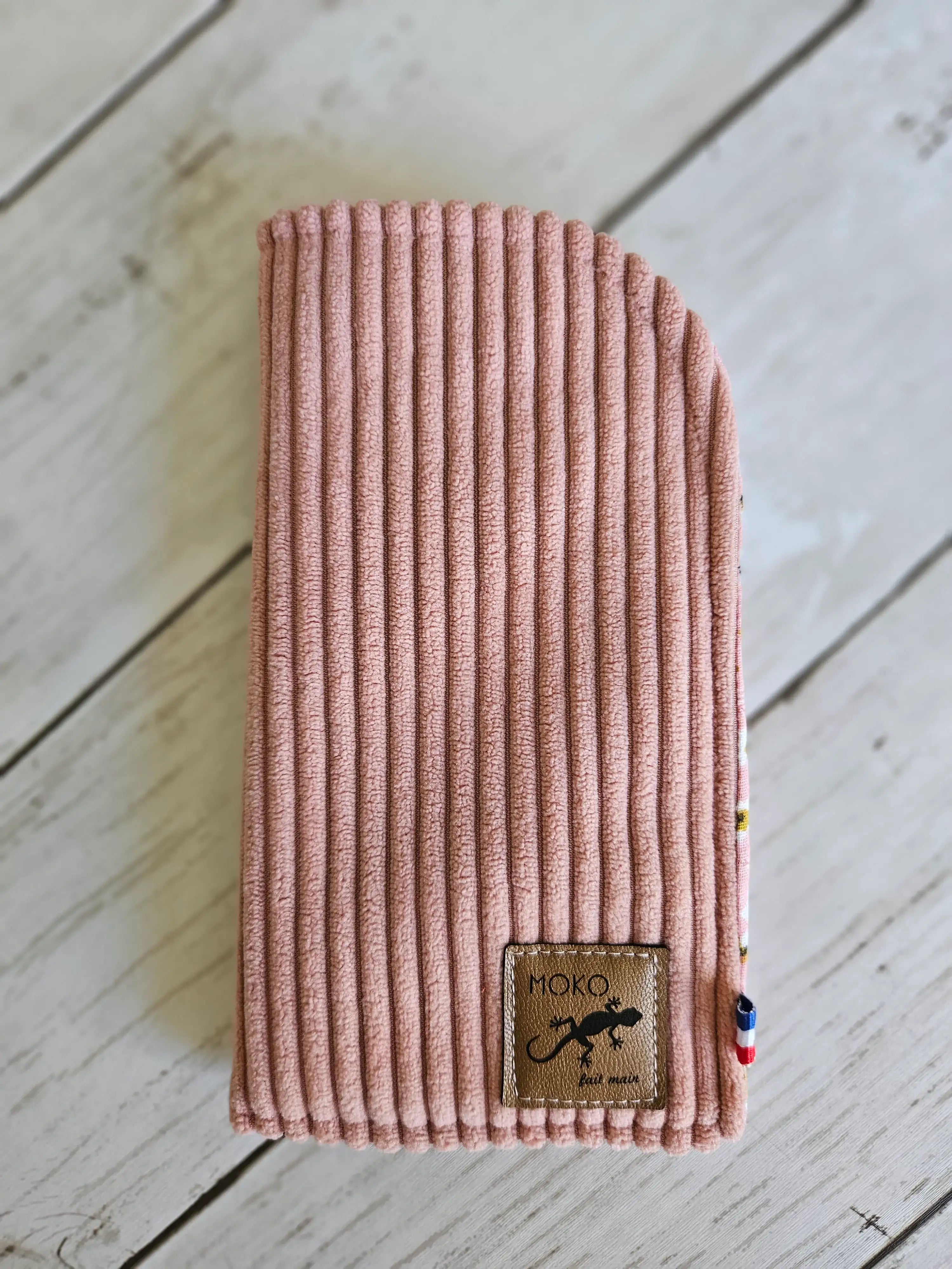 Étui à lunettes en velours côtelé rose, étiquette cuir, posé sur fond bois clair.
