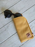 Étui à lunettes jaune côtelé en tissu sur fond bois, accessoire tendance avec lunettes de soleil.