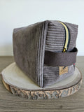 Trousse en velours côtelé gris avec fermeture éclair dorée, posée sur une tranche de bois décorative