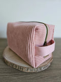 Trousse de toilette rose en velours côtelé avec fermeture éclair, posée sur rondin de bois