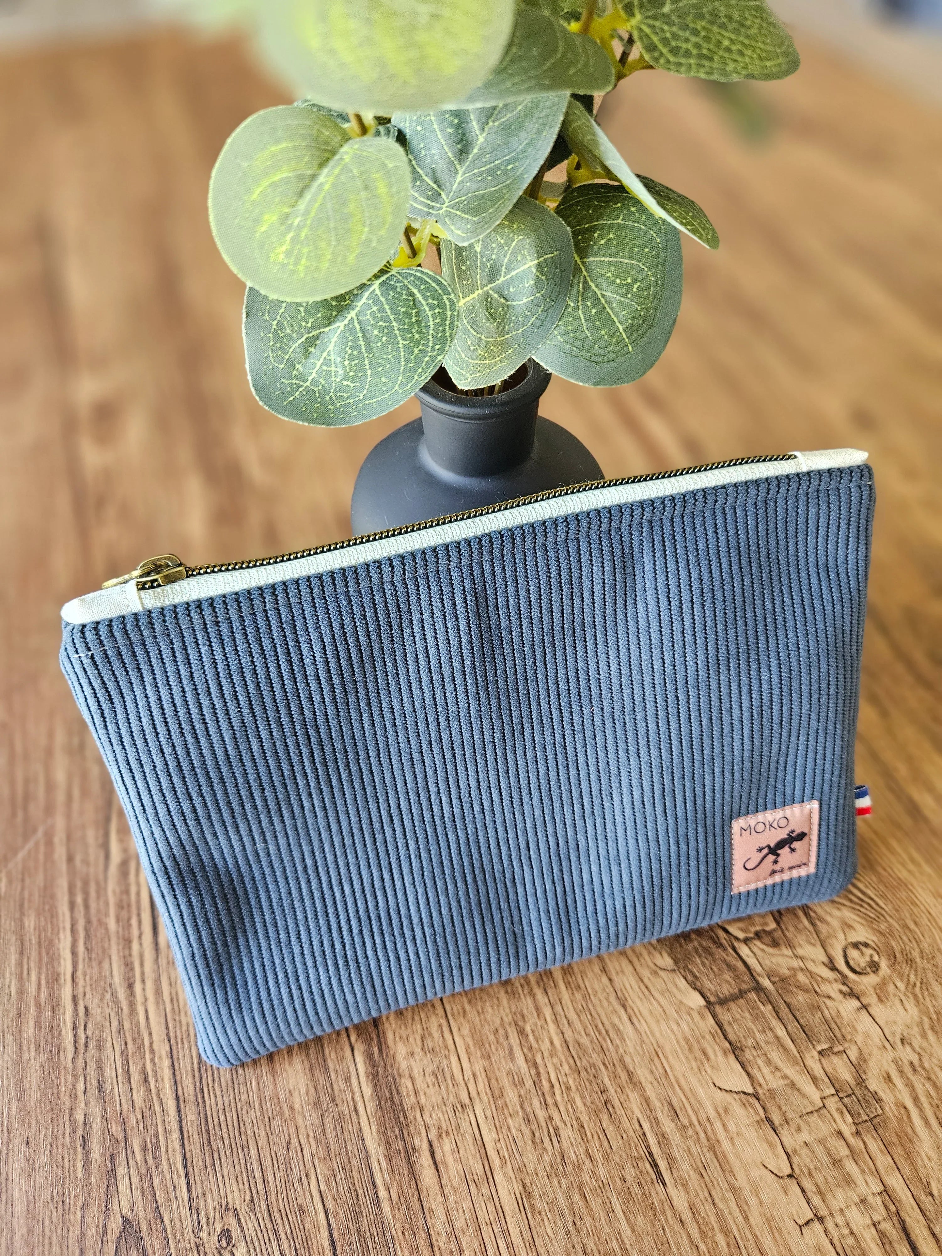 Pochette en tissu côtelé bleu avec fermeture éclair, posée sur une table en bois près d'une plante.