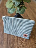 Pochette en velours côtelé gris clair avec étiquette cuir sur table en bois, plante en arrière-plan