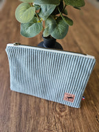 Pochette en velours côtelé gris clair avec étiquette cuir sur table en bois, plante en arrière-plan