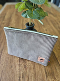 Trousse en velours côtelé gris, fermeture éclair dorée, posée sur une table en bois près d'une plante verte.