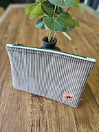 Trousse en velours côtelé gris, fermeture éclair dorée, posée sur une table en bois près d'une plante verte.