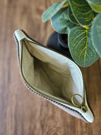 Pochette en tissu beige avec fermeture éclair dorée, posée sur une table en bois près d'une plante verte