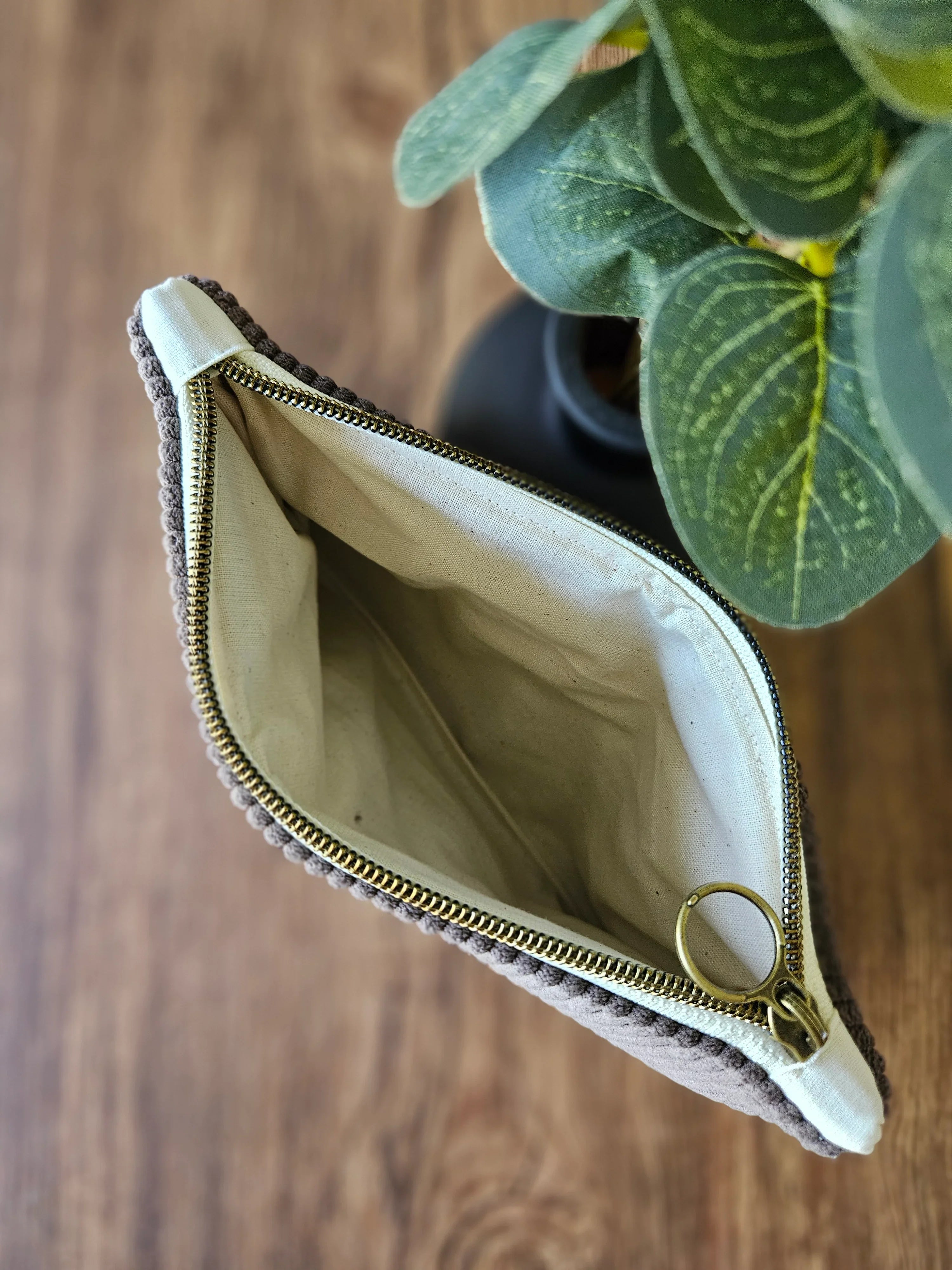Pochette en tissu beige avec fermeture éclair dorée, posée sur une table en bois près d'une plante verte