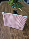 Pochette rose en velours côtelé avec fermeture éclair sur table en bois, plante décorative derrière.
