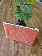 Pochette tendance en velours côtelé rose avec fermeture éclair, posée sur une table en bois, près d'une plante décorative.