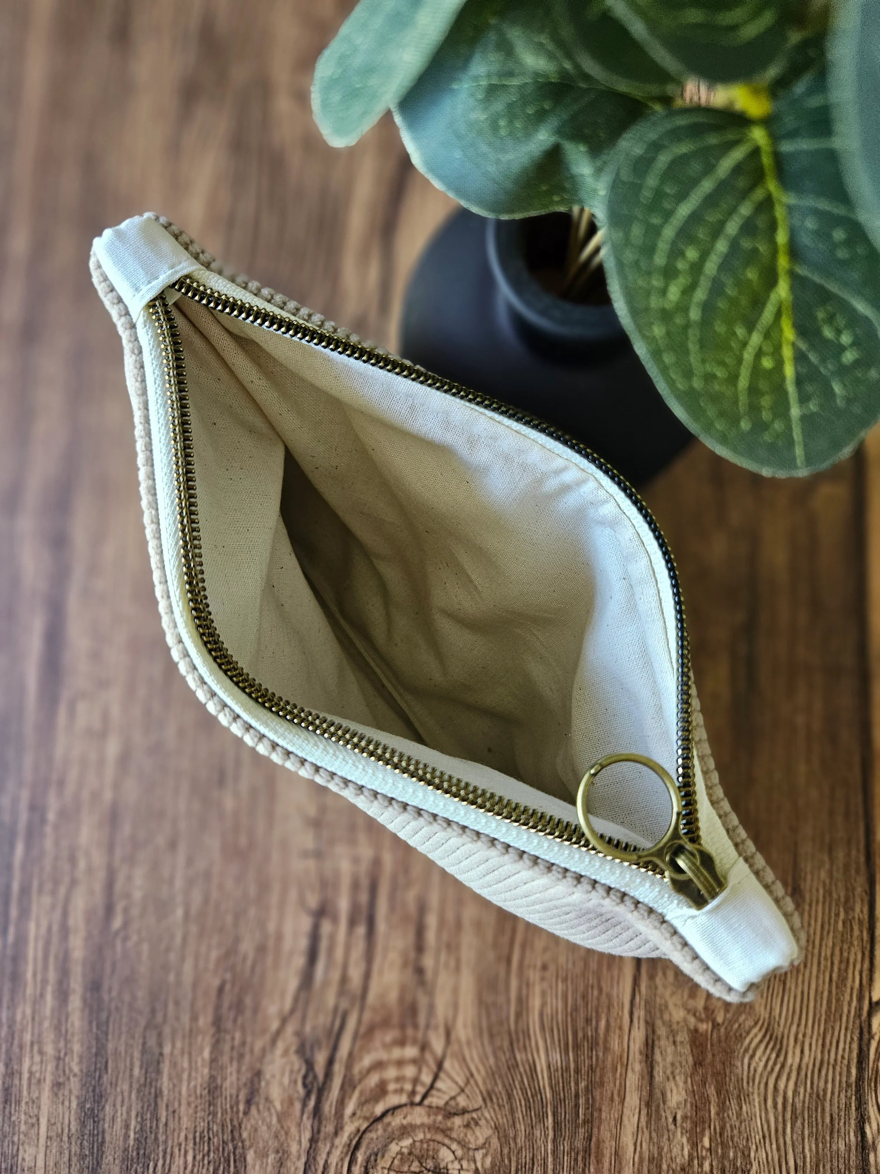 Pochette en tissu beige avec fermeture éclair dorée sur une table en bois, plante en arrière-plan